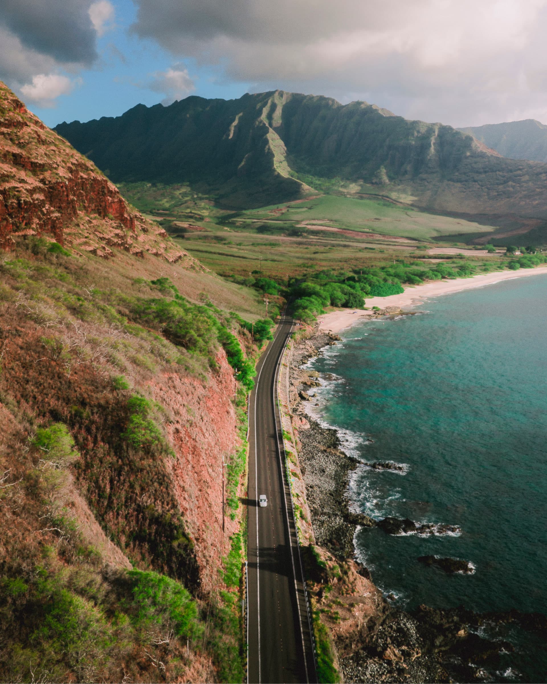 Drohnenaufnahme einer Küstenstraße auf Oahu zwischen Bergkette und türkisem Ozean